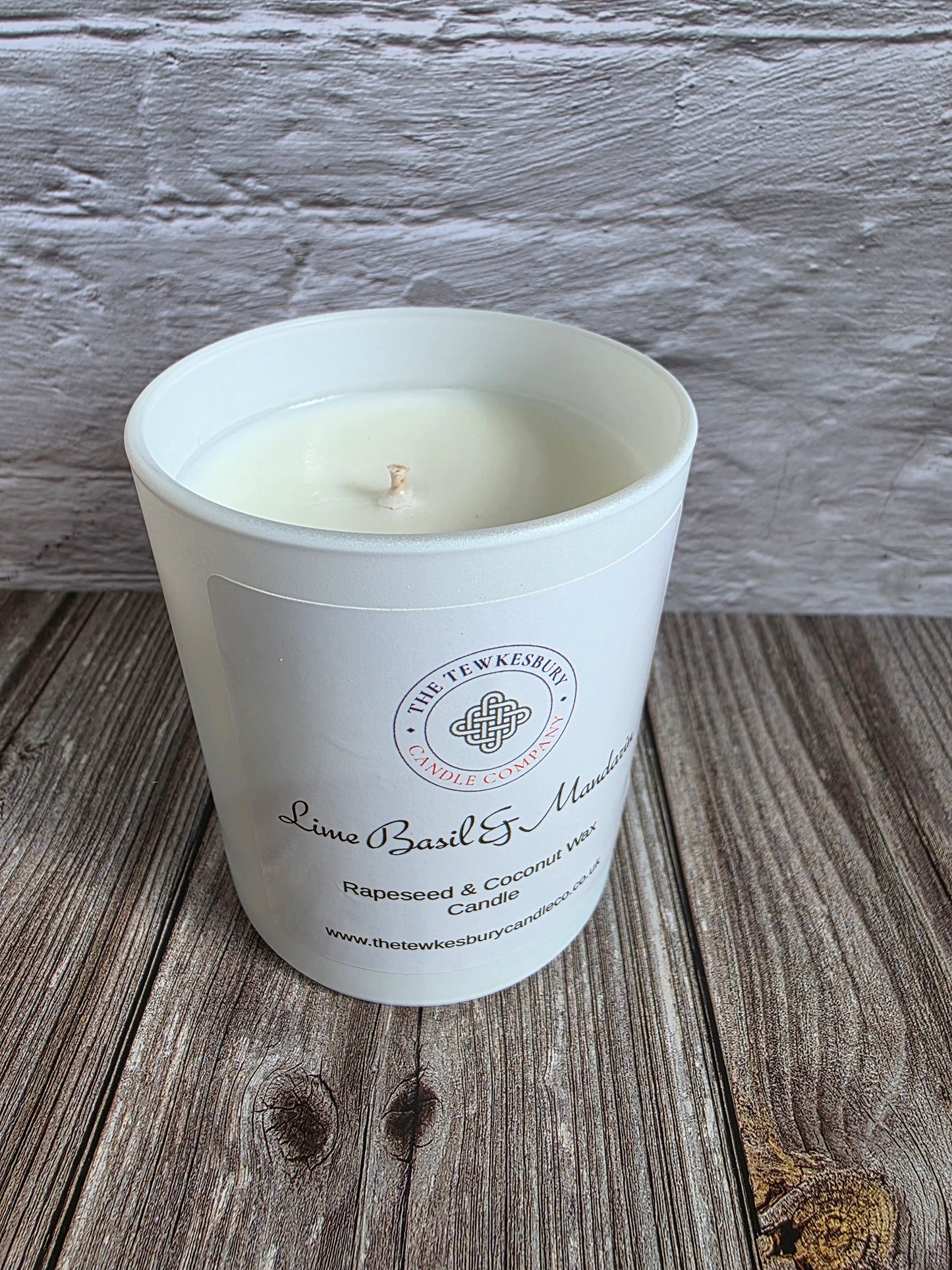 A white glass jar candle with a single wick sits on a rustic wooden surface against a white painted brick wall. The front label reads 'The Tewkesbury Candle Company' and 'Lime Basil & Mandarin'.