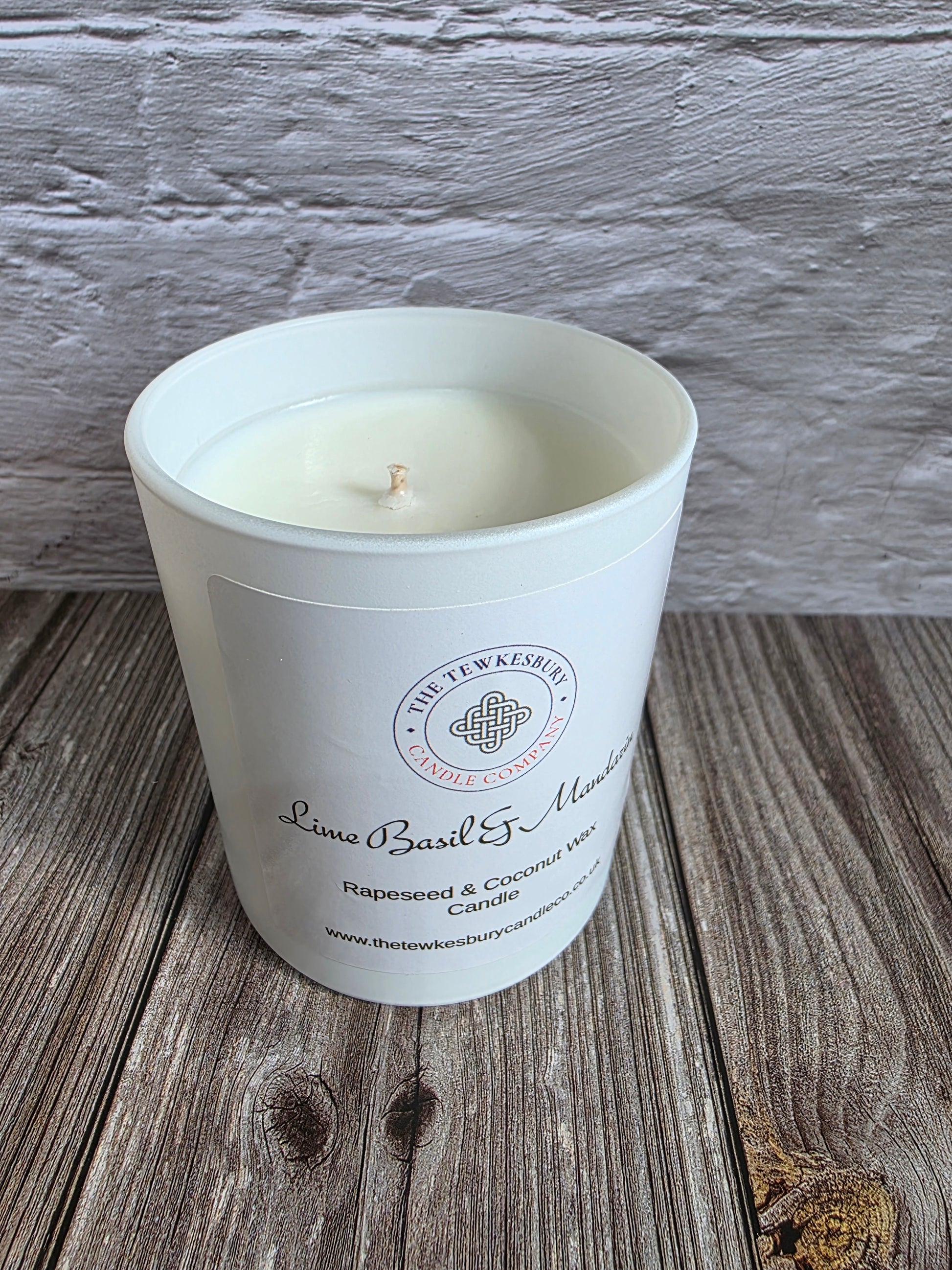 A white glass jar candle with a single wick sits on a rustic wooden surface against a white painted brick wall. The front label reads 'The Tewkesbury Candle Company' and 'Lime Basil & Mandarin'.