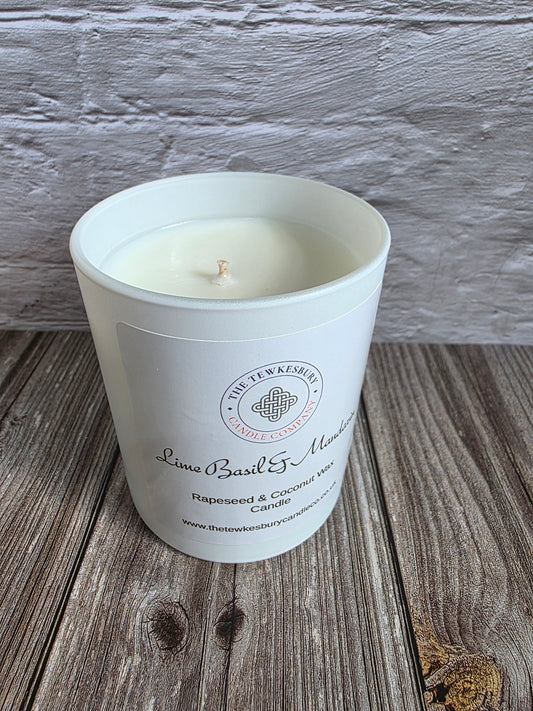 A white glass jar candle with a single wick sits on a rustic wooden surface against a white painted brick wall. The front label reads 'The Tewkesbury Candle Company' and 'Lime Basil & Mandarin'.