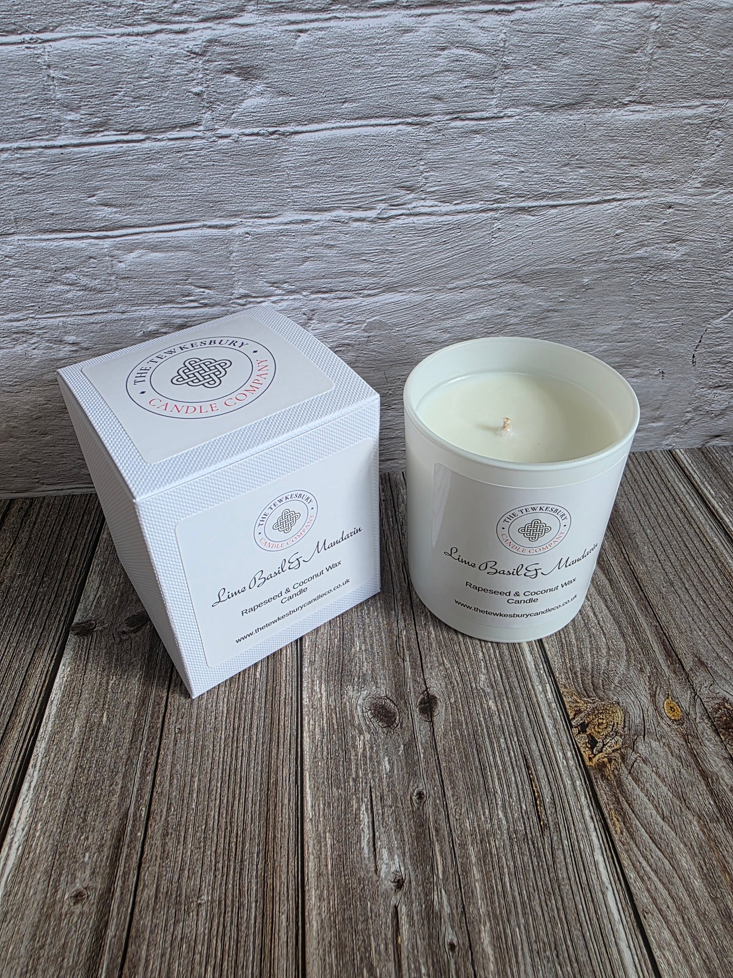 A white soy wax candle in a white glass jar sits next to its white square packaging box on a rustic wooden surface, with a white painted brick wall in the background.
