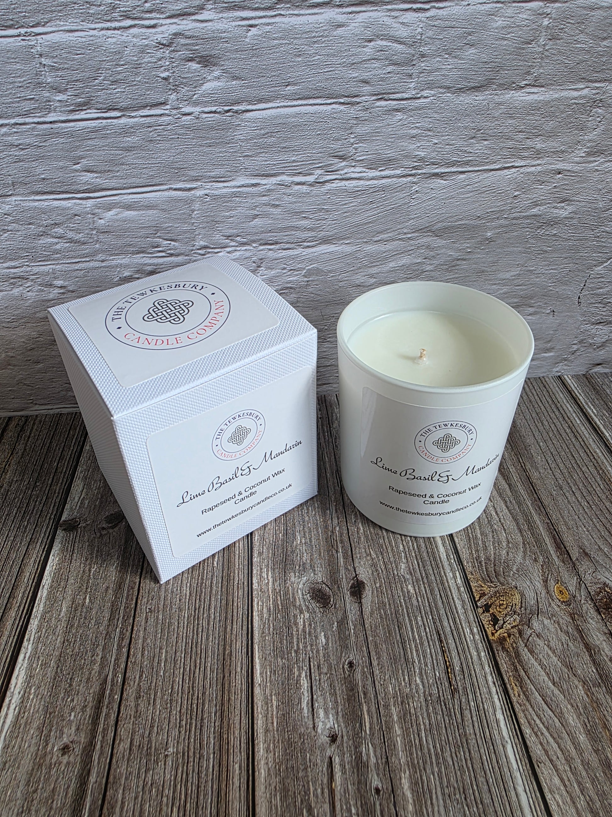 A white soy wax candle in a white glass jar sits next to its white square packaging box on a rustic wooden surface, with a white painted brick wall in the background.