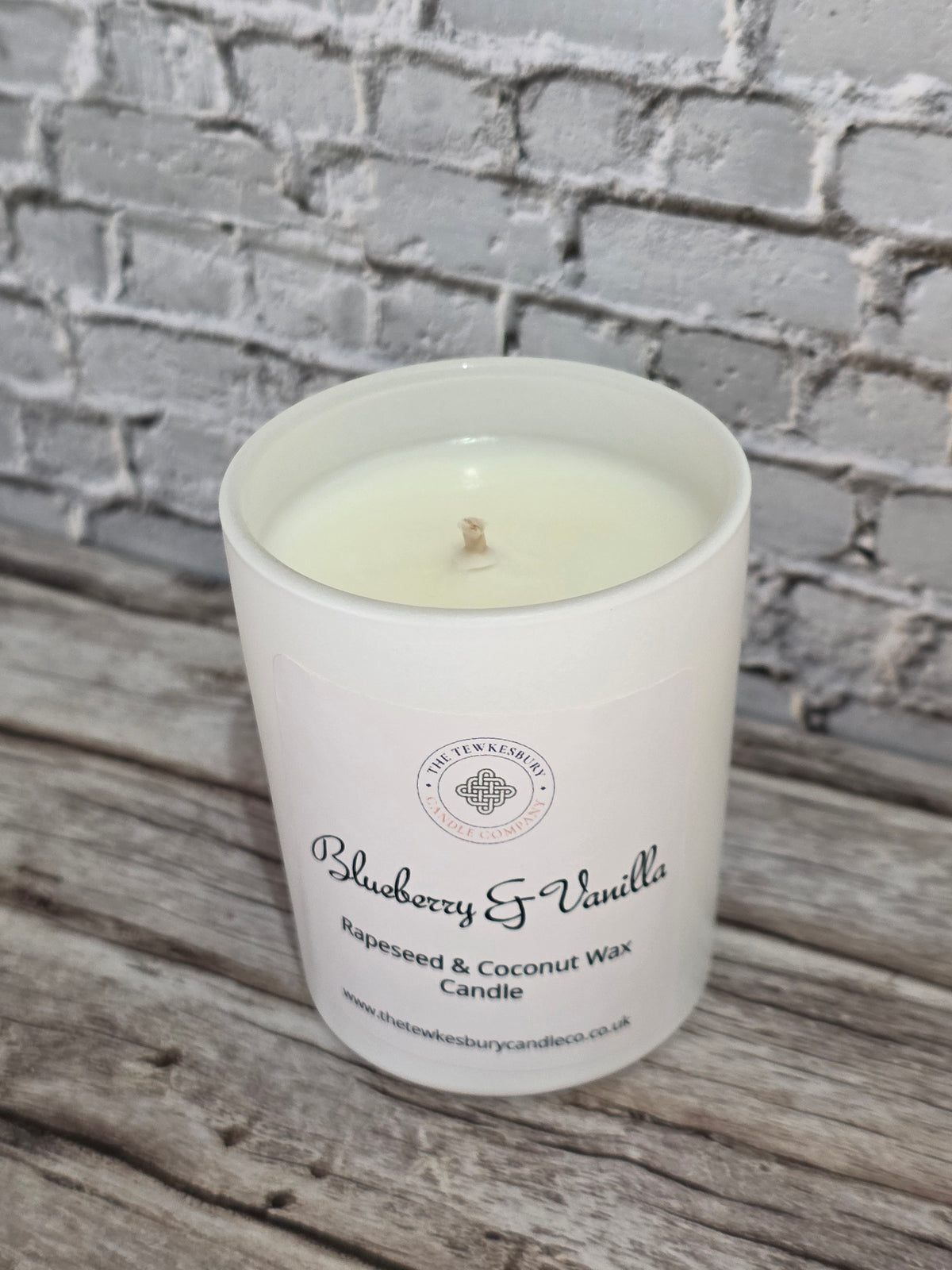 A white glass jar candle labelled 'Blueberry & Vanilla' by The Tewkesbury Candle Company, filled with creamy wax and resting on a rustic wooden surface against a gray brick wall background.