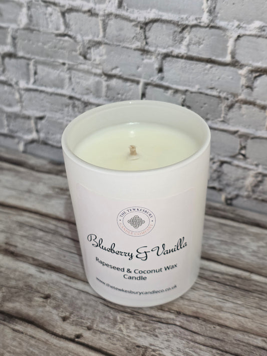 A white glass jar candle labelled 'Blueberry & Vanilla' by The Tewkesbury Candle Company, filled with creamy wax and resting on a rustic wooden surface against a gray brick wall background.