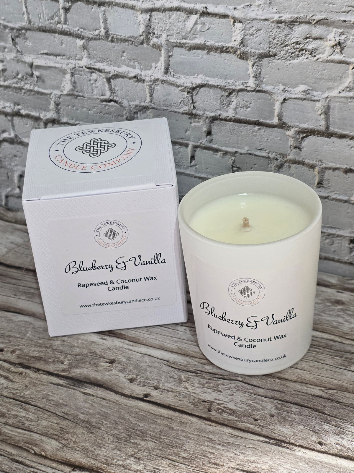 A white jar candle labeled Blueberry & Vanilla next to its matching white box, placed on a rustic wooden surface against a white brick wall background.