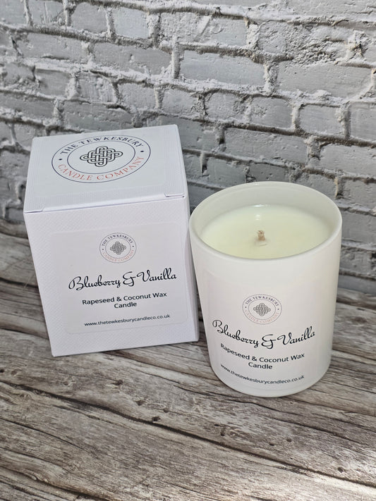 A white jar candle labeled Blueberry & Vanilla next to its matching white box, placed on a rustic wooden surface against a white brick wall background.