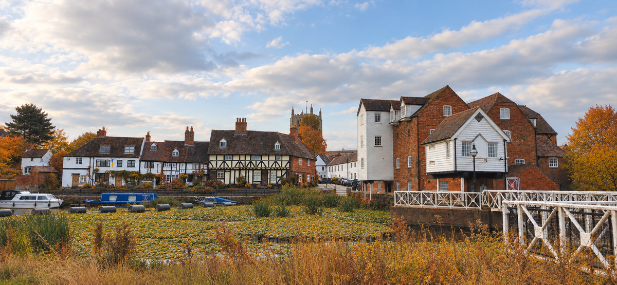 A scenic landscape photograph of a historic English village with traditional brick and timber-framed houses beside a pond under a cloudy blue sky.