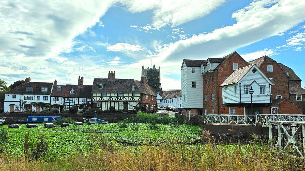A scenic landscape photograph of a historic English village with traditional half-timbered houses, a church spire, a white bridge in the foreground, and a bright blue sky with white clouds.