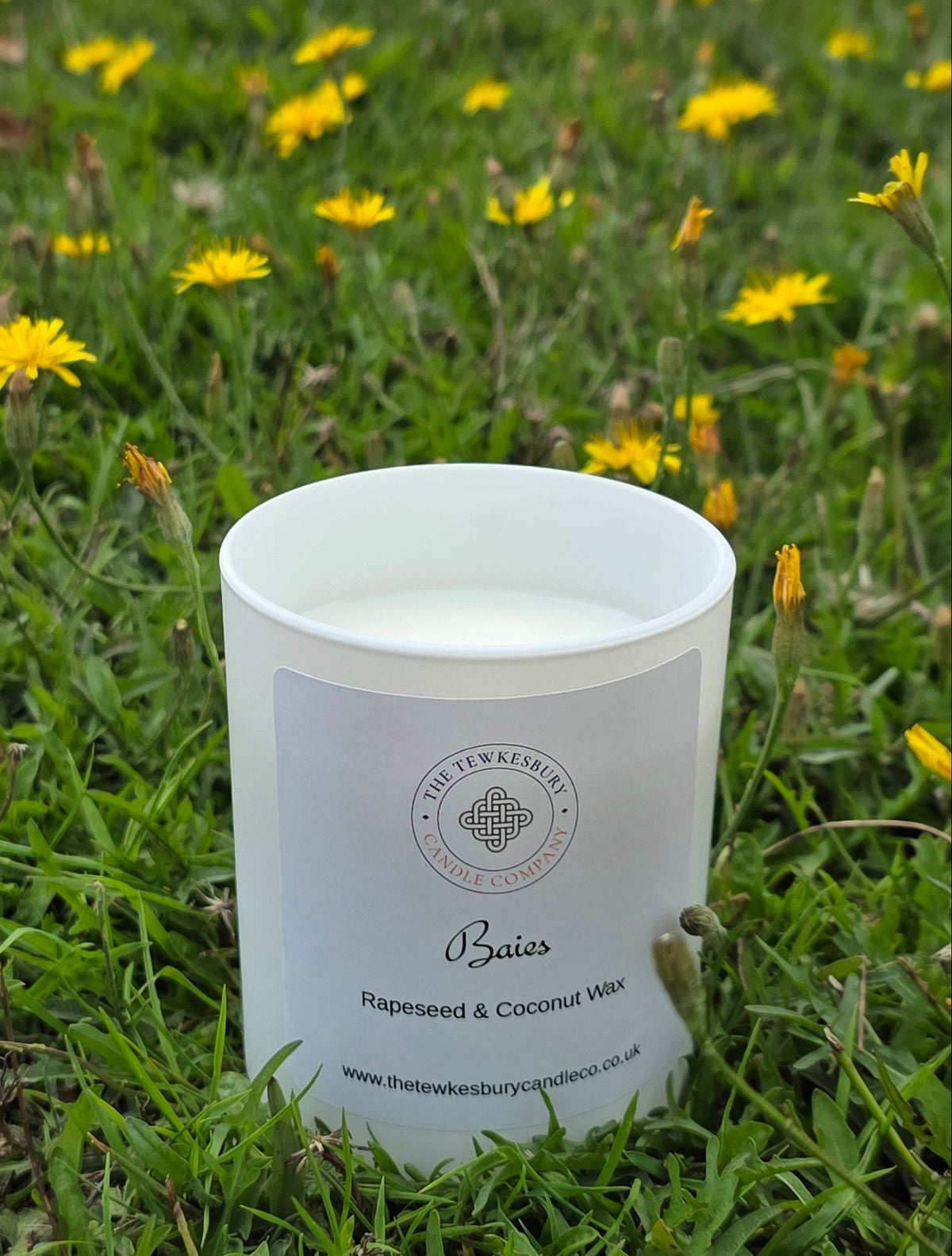 A white candle jar labeled 'Basics' by The Artisan Candle Co, placed on a green grassy field with yellow wildflowers in the background.