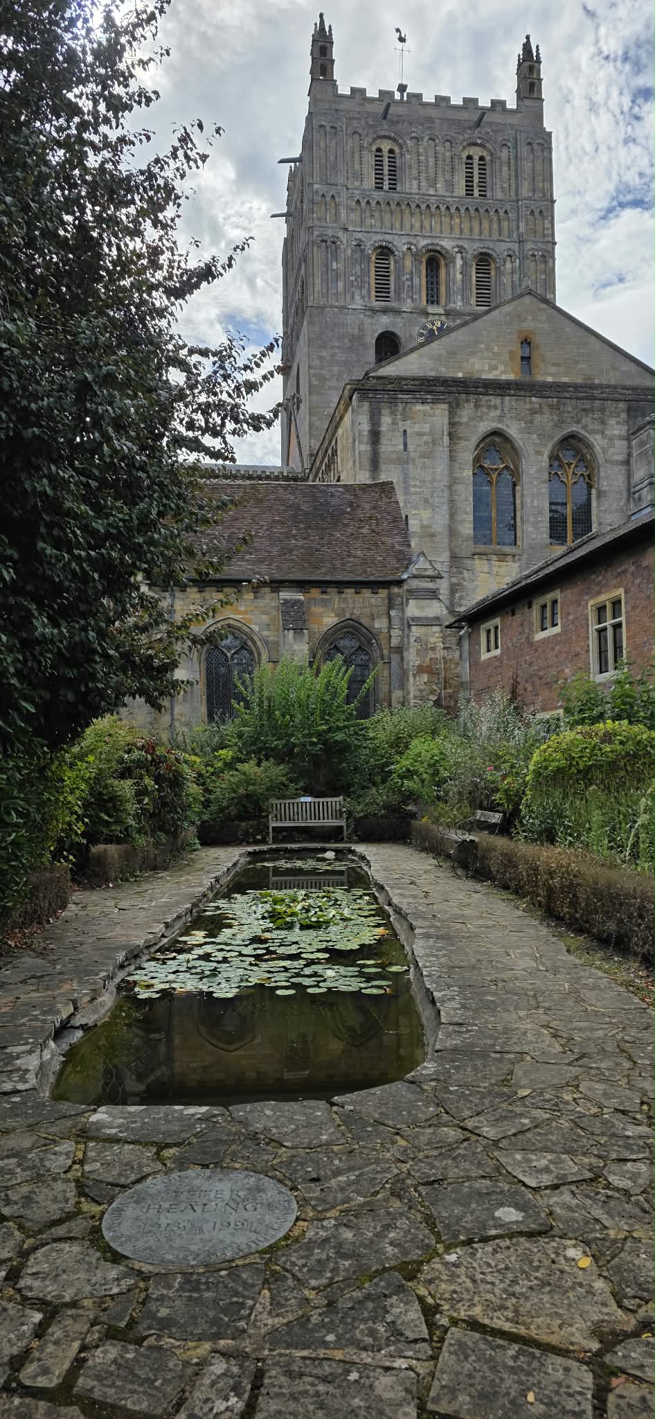 A scenic photograph of Tewkesbury Abbey, taken from the secret garden courtyard with a stone path, a small pond with lily pads, and lush greenery under a cloudy sky.
