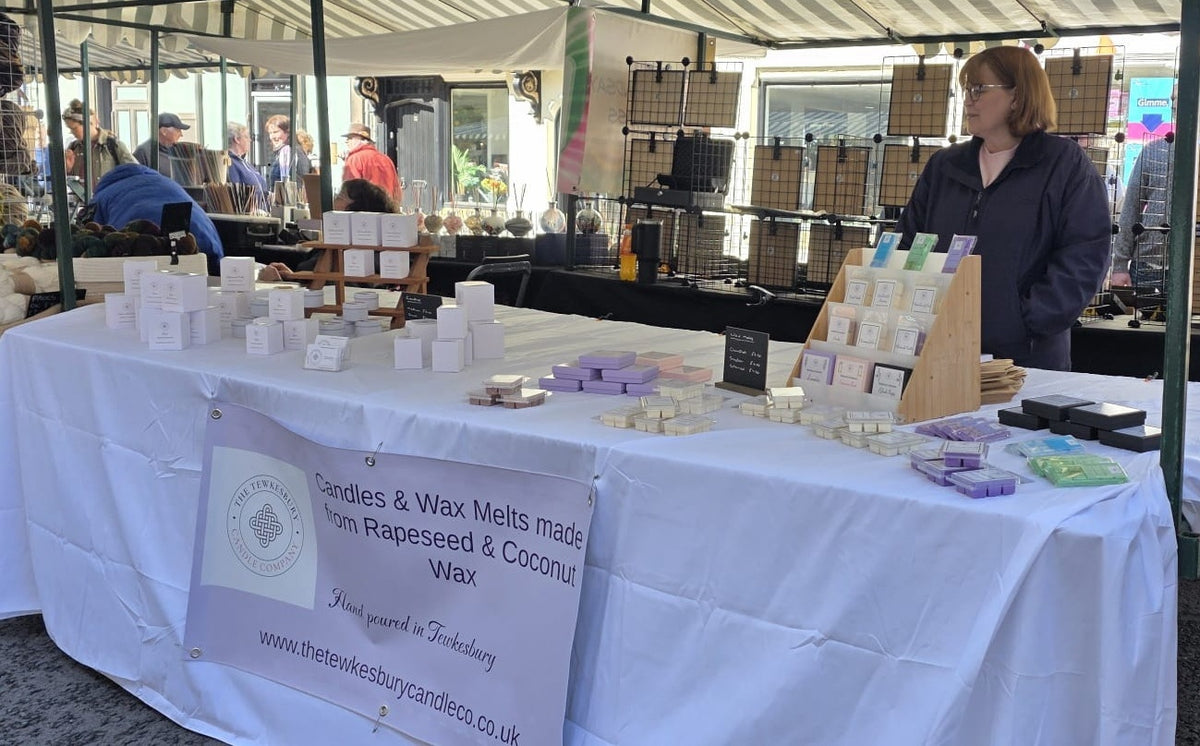A market stall display for candles and wax melts made from rapeseed and coconut wax, set up under a white canopy tent with a white tablecloth and a large sign. A woman stands behind the table, and other market items are visible in the background.